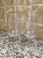 Two clear wine tasting glasses shown upright on granite countertop, etched with grapevine and grape cluster patterns.