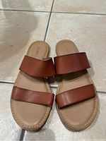 Top-down view of a pair of light brown leather sandals with two wide horizontal straps on a tiled floor.