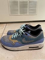 Left-side angle of the Nike Air Max sneaker showing sky blue, gold and grey textile upper, gold swoosh, blue midsole and white sole with Air Max cushioning.