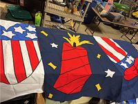 Three patriotic themed decorative flags spread out showing details with a large red firecracker on blue, red and white striped top hat, and a heart-shaped stars and stripes design