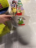 Hand holding ceramic bowl with Christmas tree design in front of Disney Snow White and holiday figures.