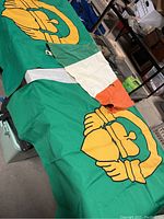 Photo showing two large green flags each with a prominent gold hand-embroidered Claudagh symbol and a smaller vintage Irish tricolor flag in green, white, and orange laid out on a table.