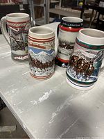 Photo of four different Budweiser beer mugs placed on a table including Clydesdale themed designs and branded logos with various scenic backgrounds