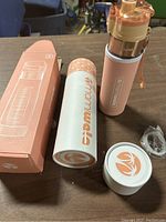 Photo showing the Mamiwata fruit infuser water bottle in pink with rose gold lid, alongside the original box, additional white container lid, and a small plastic loop accessory.
