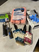 Photo showing assorted paint brushes, paint roller covers and frame, paint scraper, roller handle, and blue disposable latex gloves box.