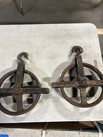 Pair of heavy duty metal pulleys side by side on a white surface, showing circular wheels and hooks.