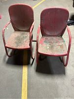 Front view of two vintage metal garden chairs side by side, showing worn red paint and visible rust and dirt on seat and backrest.
