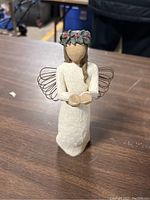 Full view of the Willow Tree Angel of Christmas Spirit figurine on table showing front with wire wings, floral crown and outstretched hands.