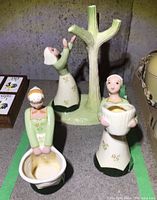 Three ceramic figurines on concrete floor. One figurine holding bowl, one reaching to tree structure, one holding round container.