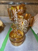 Photo showing two amber glass pitchers with white floral designs and a covered glass dish with a deer figurine lid.