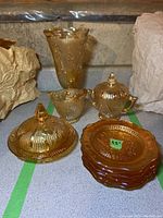 Overview of the complete amber-coloured glassware set on countertop, showing plates stacked, creamer, sugar bowl with lid, lidded dish with lid, and vase.