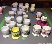 Overview of about 24 assorted egg cups in mostly white and decorated with floral and colorful designs arranged on a table.