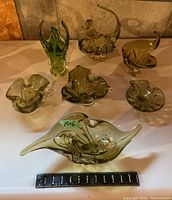 Group of seven olive green art glass pieces on white surface against cement background