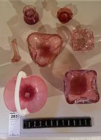 Top-down view of seven pink-toned art glass pieces including bows and a basket with a white handle beside a ruler showing size.