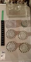Full view of approximately 12 pieces of pressed glassware including tray, three handled cups, small rectangular dish, lidded jar, and four small round pieces.