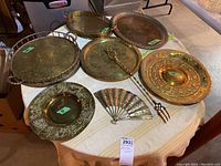 Seven metal items arranged on round table including four engraved plates, two metal trays (one warped), decorative metal fan, and large decorative fork.