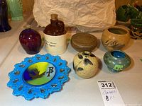 Full lot view showing various pottery pieces including vases, a jug with cork stopper, a red glazed vase, a blue glass plate with enamelled metal insert, and stacked round items.