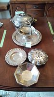 Full view of silverplate serving items on table including centerpiece with flower frog, handled compote, Wm A Rogers tray, grape design dish, Viking Plate bowl, and lidded vegetable dish.