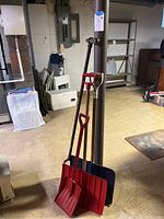 Three snow shovels and one vintage manual post hole digger standing against a basement support pole on a concrete floor.