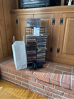 View of black metal CD rack filled with multiple CDs, white paper bag next to it on brick hearth, black Bose headphones on hearth.