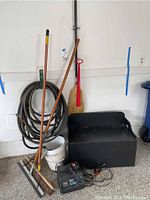 Full view of the lot showing battery charger, coiled hose, push broom, straw broom with red handle, and black painted wooden storage box containing boots.