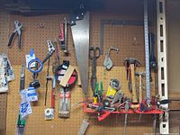 Wide shot of various hand tools and accessories hung on a brown pegboard, including saws, hammers, wrenches, and mounting tape visible.