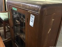 Front corner view showing glass front doors with decorative wood lattice and rounded top edges of the cabinet. Visible scratches and wear on wood finish.
