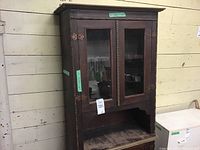 Front view of vintage wooden cabinet showing two glass-paneled upper doors with brass hinges, open shelf below, and worn wood surface.