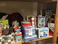 Photo showing various stacked and arranged Christmas-themed tins with colorful festive designs on a wooden shelf, including snowmen, winter scenes, and floral tins.