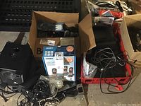 Photo showing two black Logitech satellite speakers, a black subwoofer with a circular port and cable, a box of Uniden 6.0 GHz cordless phones with packaging intact, various electrical cords, phone handsets and base units in boxes and crates.