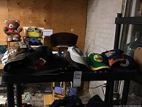 7 different hats placed on a black table including baseball caps in multiple colors and a black felt hat with feather decoration.