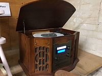 Front angled view of the wooden music center showing the open lid revealing the turntable and the black control panel with knobs, display, CD slot, and cassette deck.