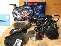 Bosch drill box, circular saw blades, automotive jumper cables, electronic tester pouch, and other components arranged together on wooden surface.