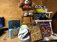 Photo showing children's rocking chair, doll high chair with teddy bear, Minion plush toy, metal lunch box, Bingo game, Disney 1000-piece puzzle, folded blanket.