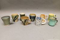 Full front view of all nine studio pottery mugs arranged in a row showing various designs and colors