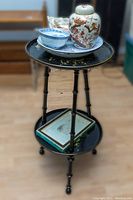 Full view of black lacquered two-tier plant stand with floral designs, holding assorted ceramic items.