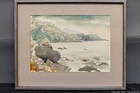 Framed watercolour painting showing a rocky coastal scene with cliffs, rocks, and water in soft muted colors.