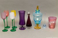Front view of multi-colored glassware including tulip wine glasses, purple goblet glass, cranberry decanter with stopper, two-toned covered candy dish, small clear stemmed glass, and pink floral glass.