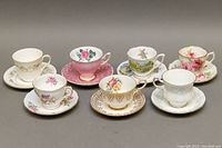 Front view of all seven teacup and saucer sets arranged in two rows showing different floral and gold rimmed patterns.