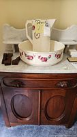 Front view of antique wooden washstand with marble top, porcelain pitcher and basin set placed on top.