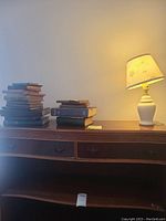 Stacked hardback books next to white ceramic lamp on wooden desk, lamp is turned on.