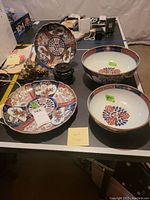 View of two large Asian nesting bowls with floral designs and two multicolored Asian decorative plates alongside wooden base and stone art piece on black table