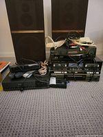 Photo showing multiple vintage electronic devices stacked on carpet including two large speakers in background, various VHS players, Fisher cassette deck, remote controls, and cables on top.