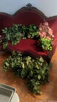 Three artificial ivy plants and one silk floral arrangement placed on and in front of a red velvet antique sofa. Easel baskets are metal and woven. Green ivy leaves with varying shades and shapes. Silk pink floral arrangement in round brass pot.