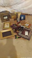 Photo showing a group of 13 assorted frames of different sizes and finishes lying on a basement floor, some with artwork or placeholder images inside, showcasing variety in styles and material.