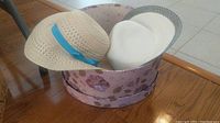 Two woven hats in a round floral decorative hat box on a wooden table, one featuring a blue ribbon.