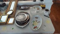 Photo showing assorted rectangular framed mirrors, silver plate trays stacked, a ceramic oval tray with floral and fruit decoration, and clear glass bowls and trays arranged on a table.