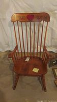 Front view of a wooden spindle back rocking chair with a hand-painted red heart and floral design on the top rail, showing the chair seat and armrests.