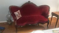 Front view of carved wood velvet-look settee with floral decorative pillow on a wood floor near a lamp and table.