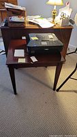 Angled front view of wooden side table with paperwork and printer on top. Shows table legs, surface wear, and overall shape.